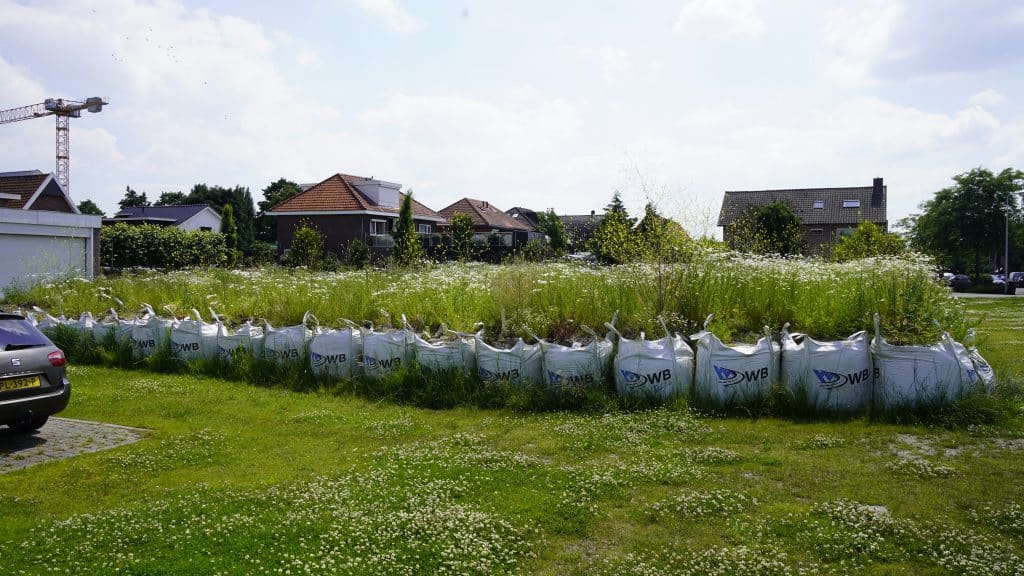 Een foto van hoe het terrein van de Beukenhof er in juli 2024 bij ligt. Een groen grasveld met daarop een verzameling van witte zandzakken waar groene planten met witte kopjes uitsteken.