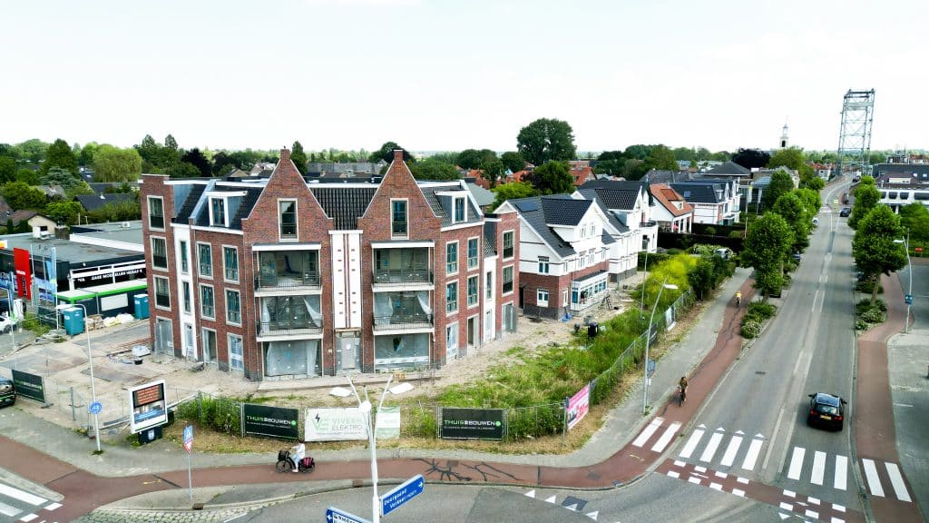 Foto vanuit de lucht waarop links het appartementengebouw te zien is van het nieuwbouwproject Kerkweg-Oost 214-216. Rechts loop de Kerkweg-Oost met een auto die richting de Hefbrug rijdt. Achter het appartementengebouw zie je de andere woningen die bij dit project gebouwd zijn en op de achtergrond ook de villas op nummer 222. 