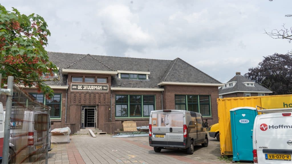 Een groot bakstenen school in renovatie, met een de nieuwe naam op het bord boven de ingang: ‘DE STUURMAN’. Voor de oude school, op het voormalige speelterrein liggen bouwmaterialen en staan werkbussen. Foto is genomen vanaf de ingang op de Oranjelaan.