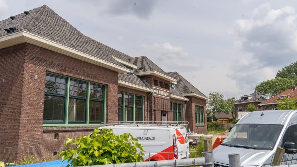 Een groot bakstenen school in renovatie, met een de nieuwe naam op het bord boven de ingang: ‘DE STUURMAN’. Voor de oude school, op het voormalige speelterrein liggen bouwmaterialen en staan werkbussen. Foto is genomen vanaf de ingang op de Oranjelaan.