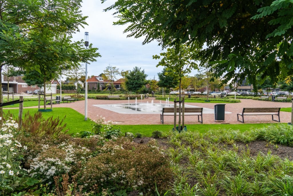 Een foto van het Beukenhofplein voor het gemeentehuis. De foto is vanaf de Kerkweg-Oost genomen. Er is veel groen te zien en het plein is omringd met bankjes. In het midden van het plein zijn er verschillende waterstralen van een fontein te zien.