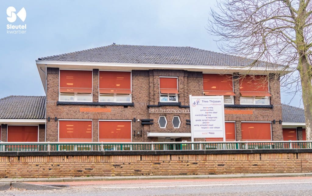 Een bakstenen schoolgebouw van twee verdiepingen met rode luiken, met een bord ervoor met de naam van de school: ‘Theo Thijssenschool’. Op het bord staan verder de kernwaarden van de school. De foto is genomen vanaf de Kerkweg-Oost verhoging richting de Hefbrug.