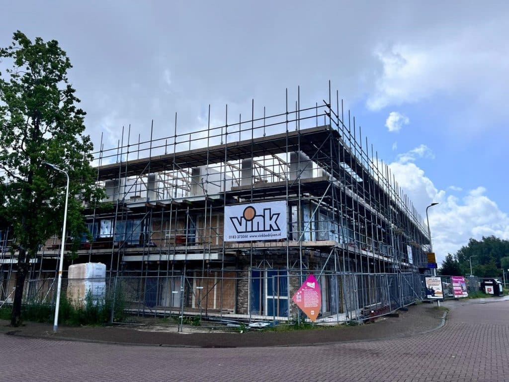 Het gebouw in aanbouw is omgeven door steigers, de foto is genomen vanaf de kruising op de Stationstraat en de Jan Dorrekenskade. Er zijn al twee verdiepingen klaar en de derde is in aanbouw. Een spandoek met de tekst ‘Vink’ is prominent op de constructie geplaatst. De lucht is gedeeltelijk bewolkt en er staat een boom aan de linkerkant van de foto.