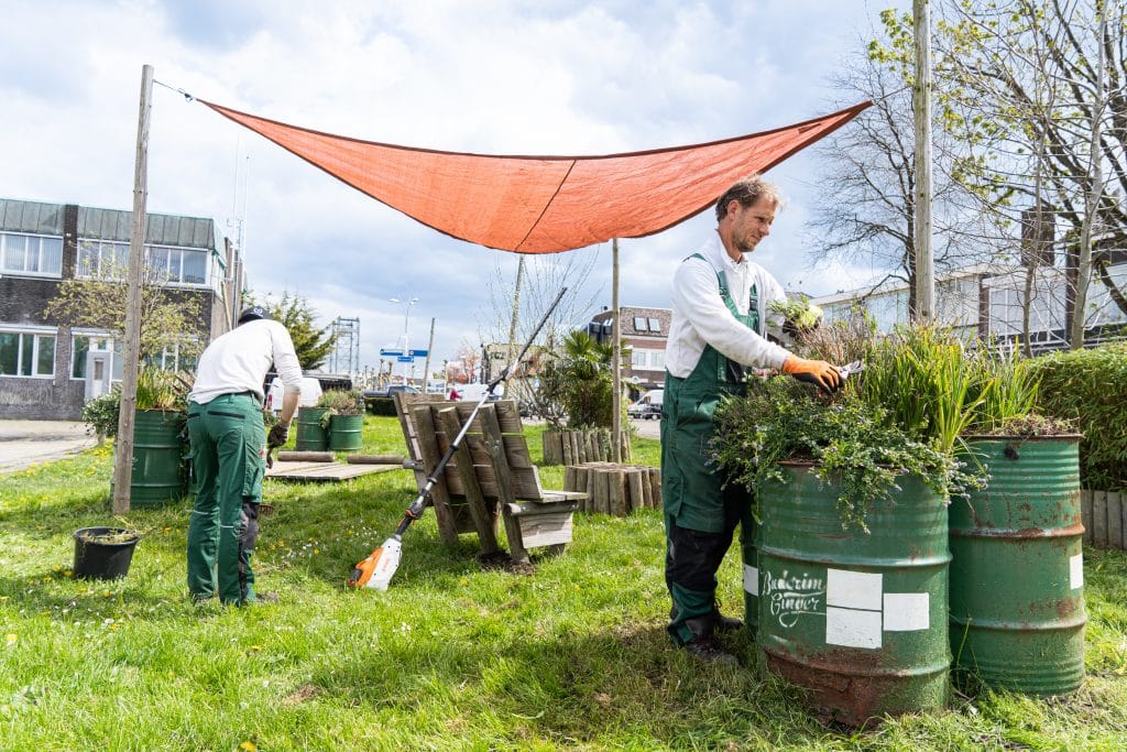 Een foto van twee tuinmannen die het stadsstrand op de Passage klaar maken. Zij knippen planten en harken het gras. Het stadsstrand bestaat uit houten banken en een oranje doek is daarboven gespannen.