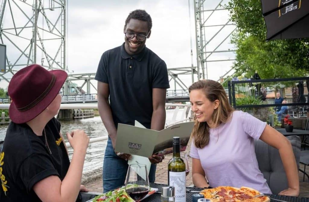 Twee personen op een terras aan de Nesse. Zij worden een menukaart voorgehouden door de serveerder. Op de achtergrond is de Hefbrug te zien. 