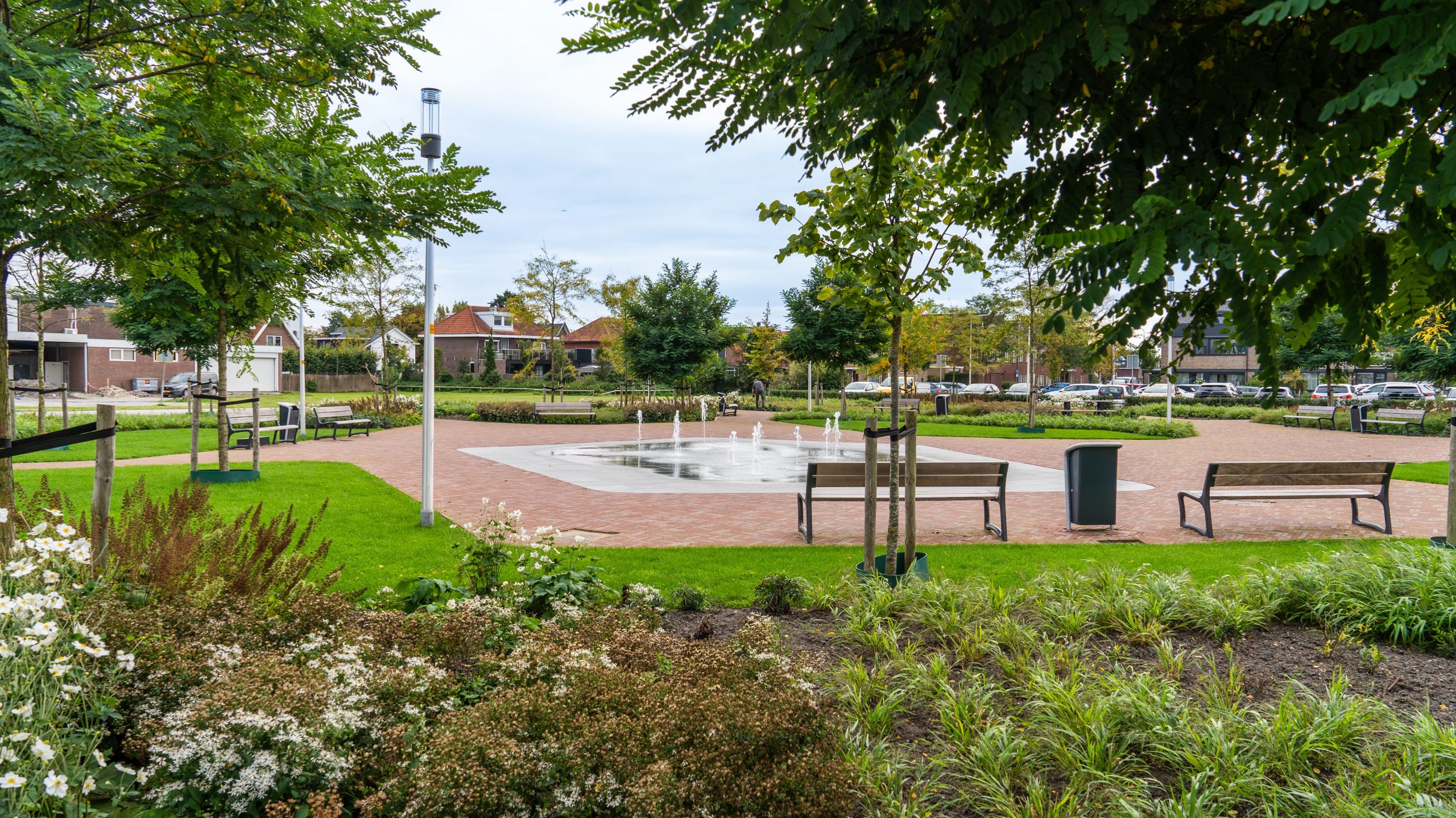 Een foto van het Beukenhofplein voor het gemeentehuis. De foto is vanaf de Kerkweg-Oost genomen. Er is veel groen te zien en het plein is omringd met bankjes. In het midden van het plein zijn er verschillende waterstralen van een fontein te zien. 