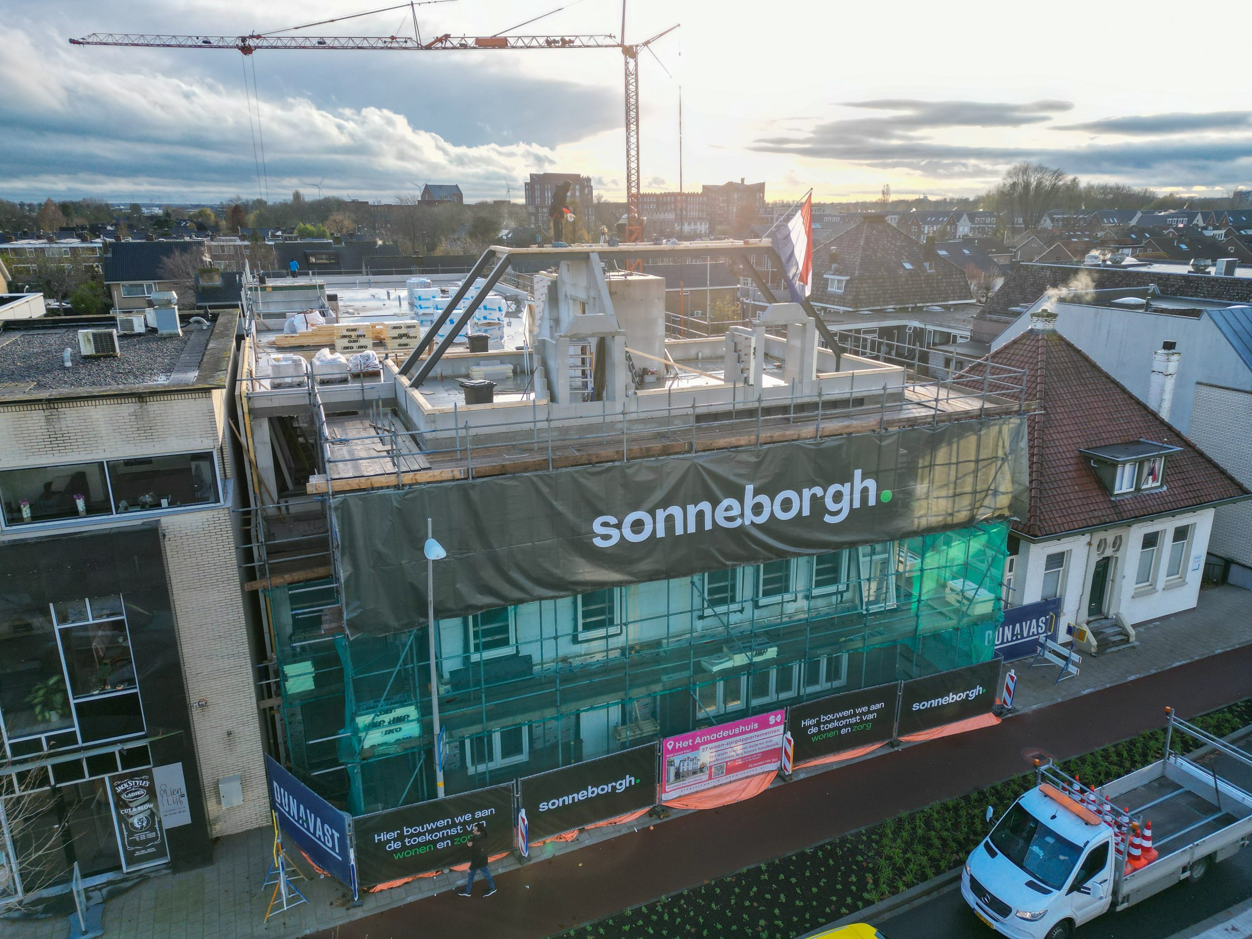 Het amadeushuis in aanbouw, het hoogste punt is bereikt. De vlag hangt uit. Je ziet de betonnen muren. en het begin van het metselwerk onderaan. Er staat een hijskraan boven